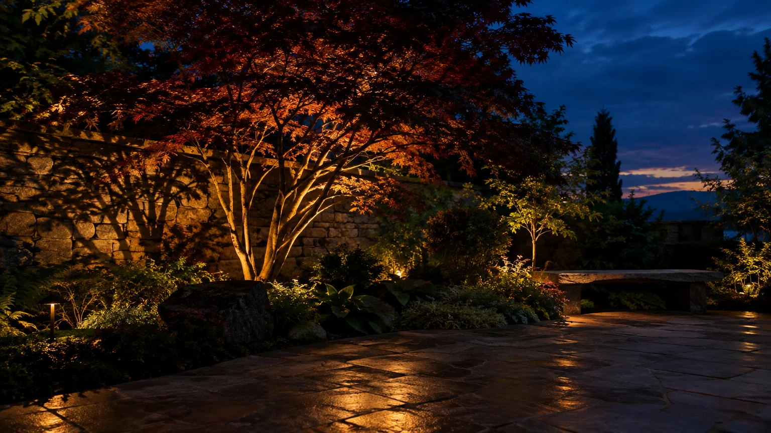 Warm uplighting illuminates a Japanese maple casting shadows on a stone wall beside a flagstone patio at twilight.